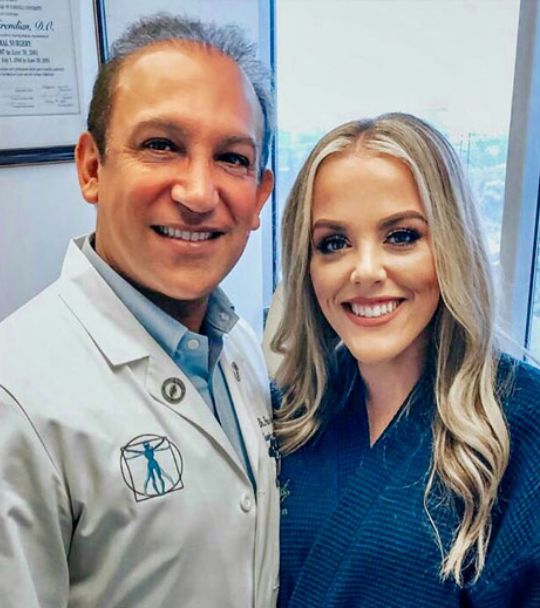Smiling man in a white medical coat and a smiling woman stand together in a clinical office setting, posing for a photo near a window.