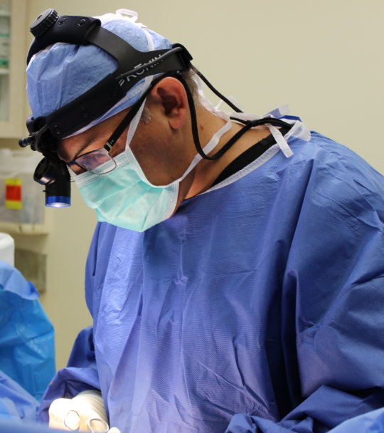 A surgeon in scrubs and a headlamp focuses intently on a procedure.