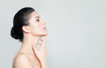 A profile of a woman with dark hair in a bun, touching her jawline with her right hand against a plain background.