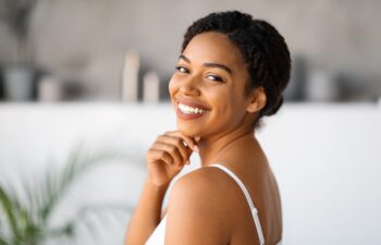 Woman with dark braided hair, wearing a white tank top, smiles while looking over her shoulder in a bright indoor setting. A green plant is visible in the background.