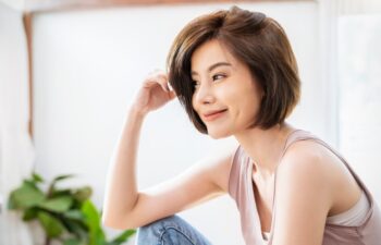 Woman with short brown hair and light skin sits indoors, smiling and looking to the side. She is wearing a sleeveless top. A plant is visible in the background.