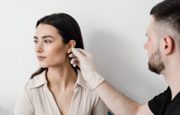 A healthcare professional wearing gloves examines a woman's ear while she looks away, against a plain white background.